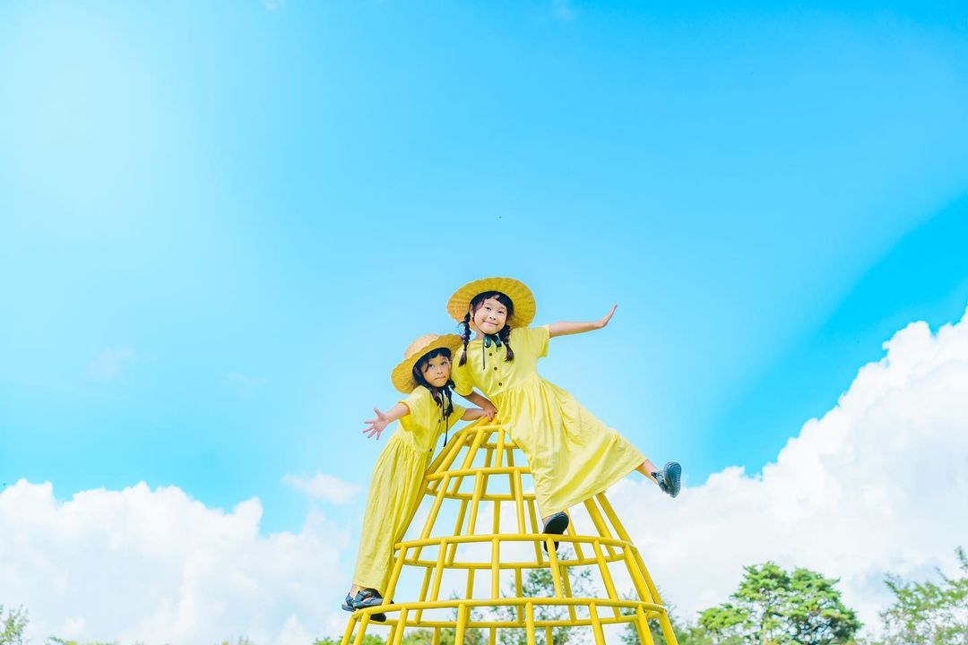 「もくもく雲」 もくもくした雲を背景に公園の黄色い遊具の上で楽しそうにポーズを取る黄色の服を着た2人の少女の写真