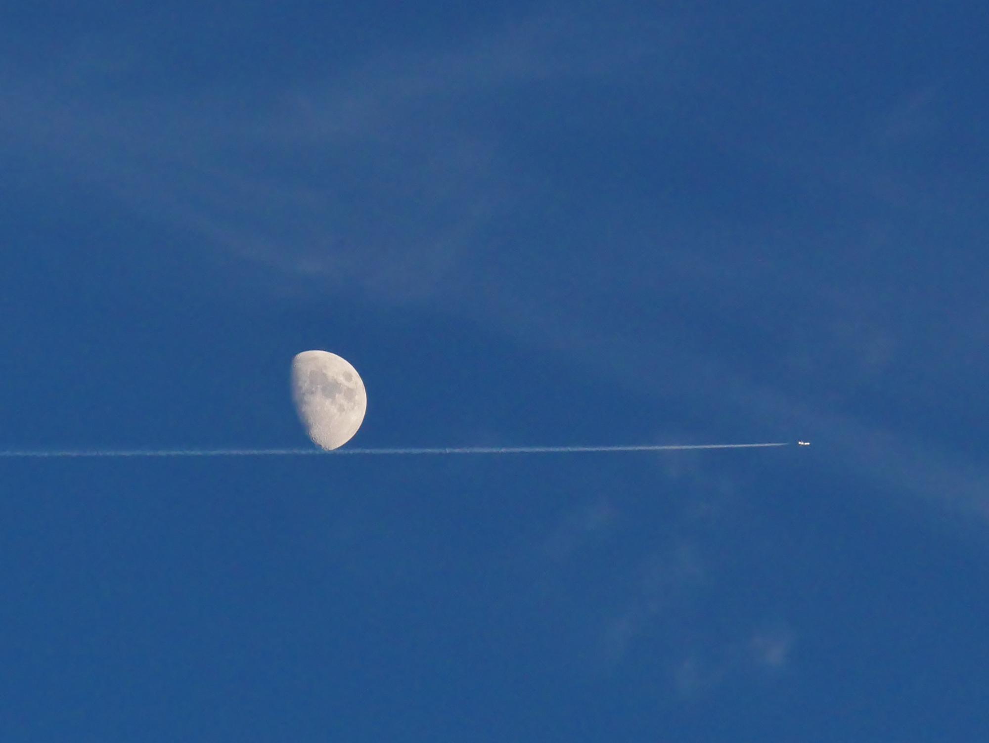 月が映える空に一筋の飛行機雲と飛行機が飛んでいる様子を捉えた写真