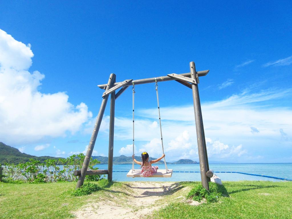 青空のもと海沿いの崖に設置されている木製のブランコをこいでいる女の子を捉えた写真
