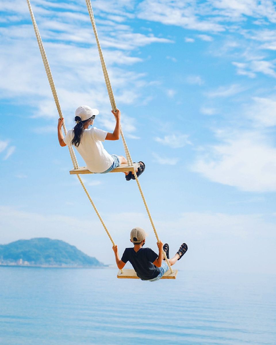 青い空と青い海に向かって二段式のブランコをこいでいる男の子と女の子の様子を捉えた写真