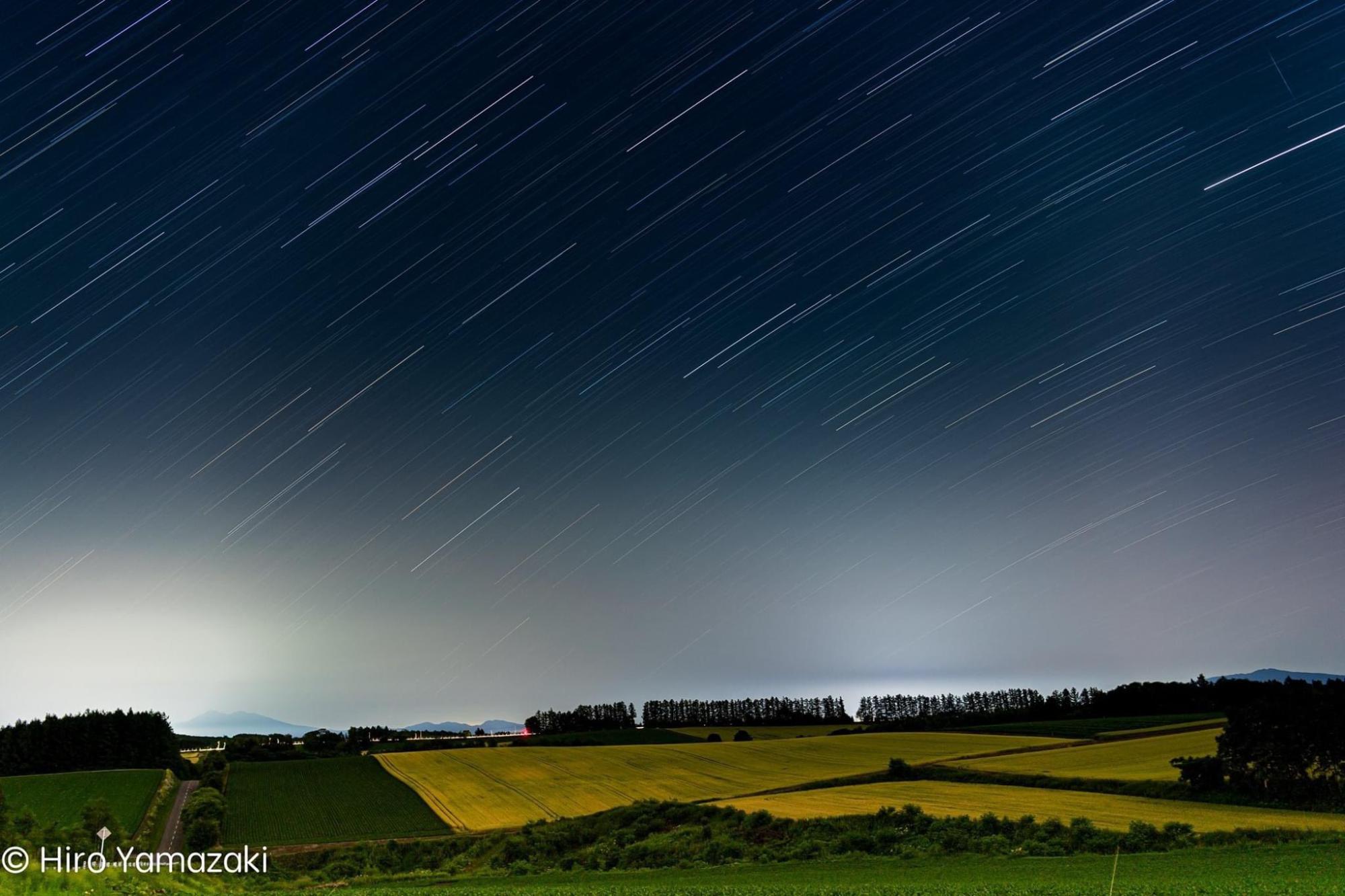 丘の上に広がる一面の闇夜に星の光が線のように降り注いでいる様子を捉えた写真