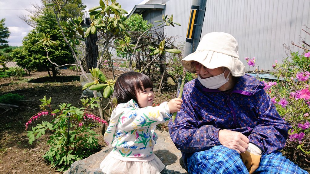 庭の石に座っているお婆さんと、アスパラガスを渡そうとしている女の子を捉えた写真
