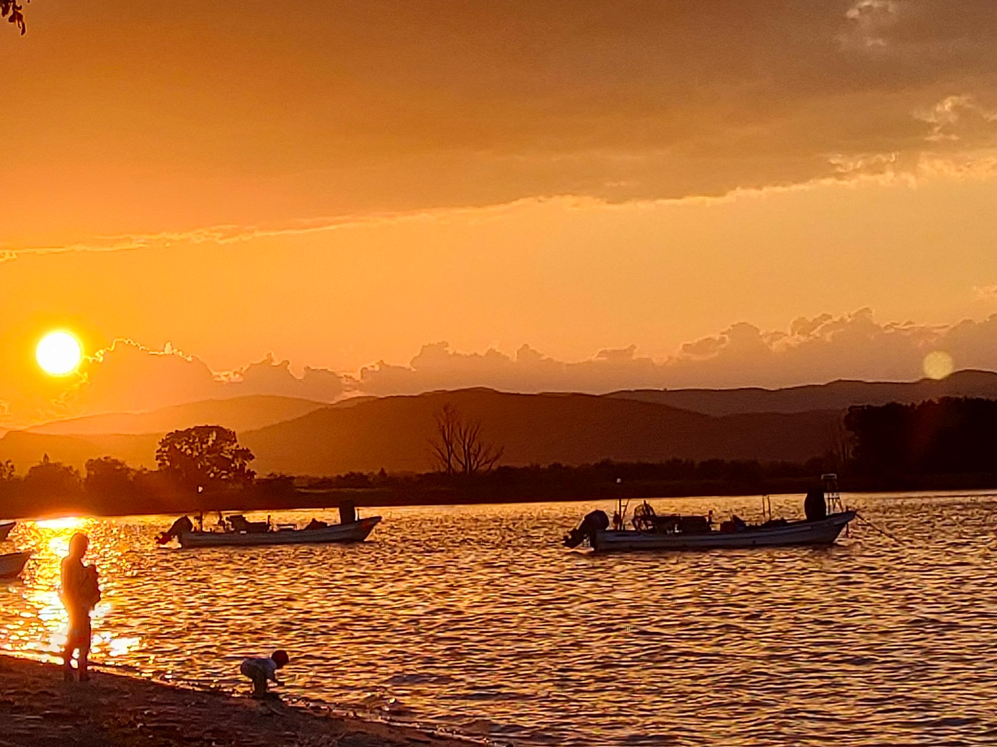 湖畔で子どもを抱っこしている女性や湖を行く二艘の船が夕日に照らされてシルエットになっている様子を捉えた写真