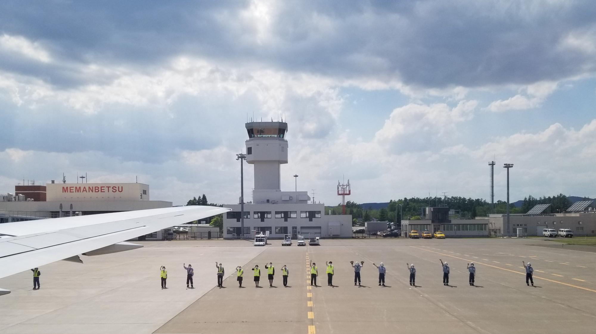 飛行機の窓から見える右翼と滑走路で横一列になって大きく手を降っている空港のスタッフたちの様子の写真
