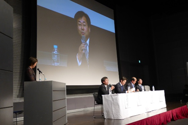 ステージ上で会見に臨んでいるスーツ姿の4人の男性と発言している男性の様子を映すプロジェクターの映像の写真