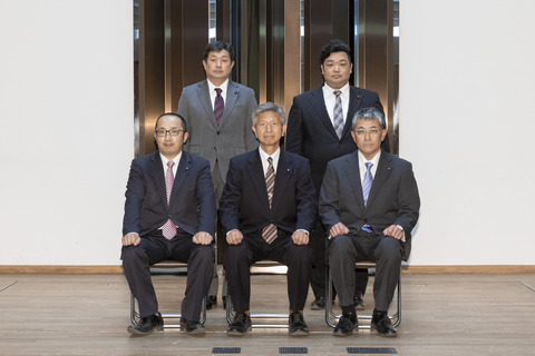 黒いスーツに茶色のストライプのネクタイをしめた男性を真ん中に、前3名が座り後ろ2名が立ち正面を向いている写真