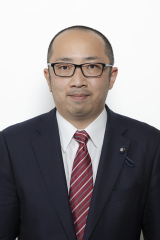 黒いスーツに赤色のストライプのネクタイをしめて黒縁のメガネをかけている上地史隆さんが正面を向いている写真