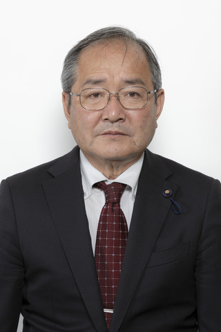 黒いスーツに赤い格子柄のネクタイをしめてメガネをかけた原本哲己さんが正面を向いている写真