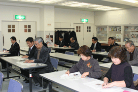 長机の席に2人ずつ座り書類を見ながら報告を聞いている様子の町民の写真