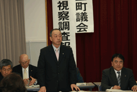 座席の前に立ち報告する様子の男性とそれを聞いている3名の関係者の写真