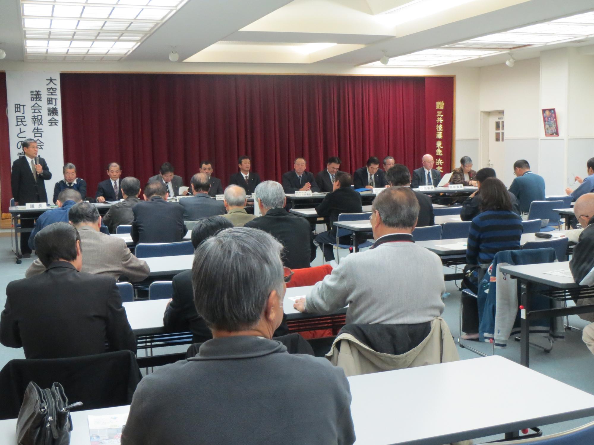 会議場で「大空町議会、議会報告会」と書かれた看板の前で発言する男性を注視する議会関係者と町民の写真。