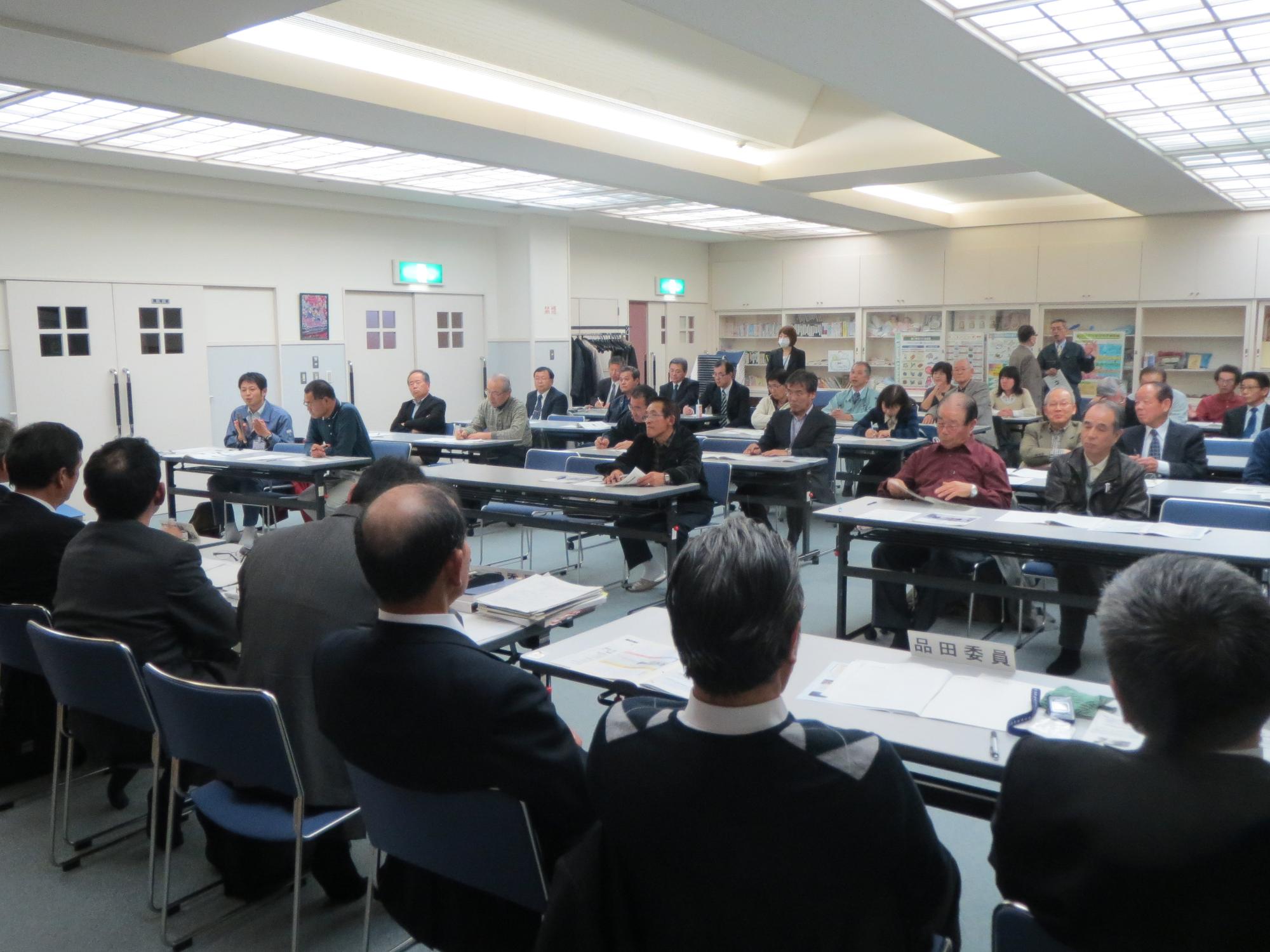 椅子と机の並んだ会議室で報告会会議に参加する町民と向かい合う議会関係者の写真。