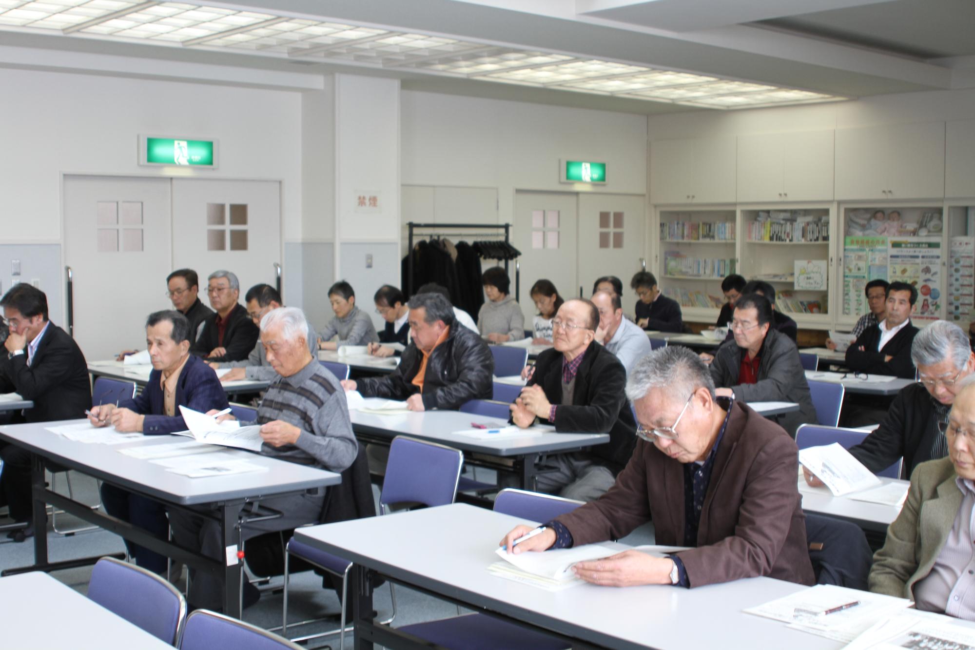 椅子と机の並んだ会議室でそれぞれ着席し報告会に参加する大勢の町民の写真。