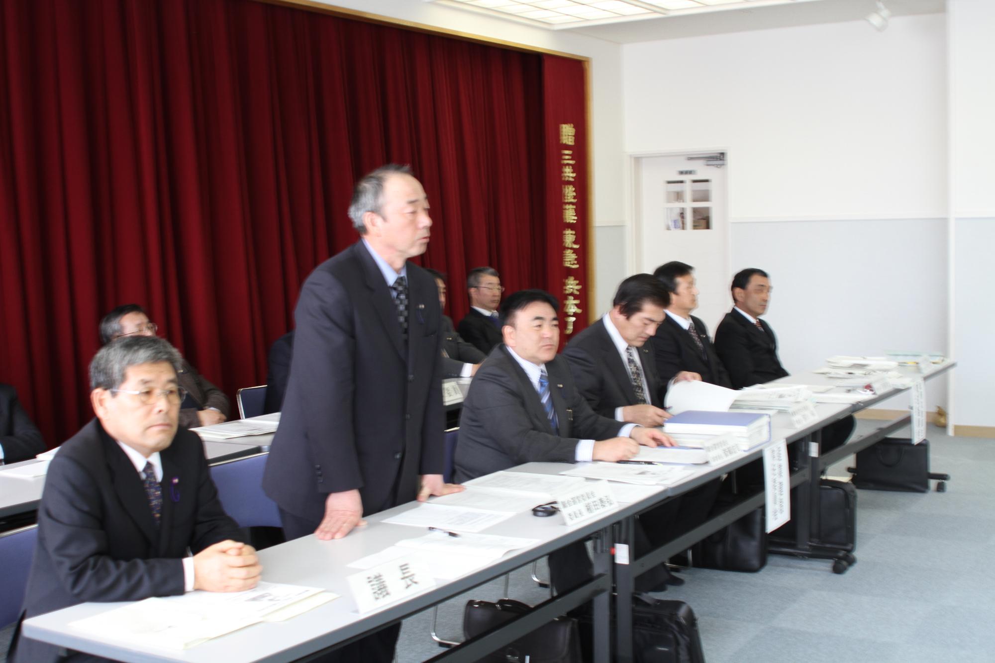会議場で机の前に起立して発言する男性とそれを注視する議会関係者の写真。