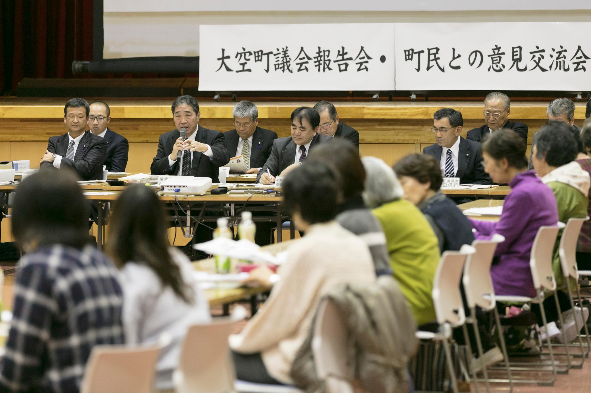 「大空町議会報告会・町民との意見交流会」と書かれた白い横断幕前の2列に並んだ長机にスーツ姿の複数の男性が座り、その中の1人がマイクを持ち周りに座っている町民の方たちに向けて話している様子の写真