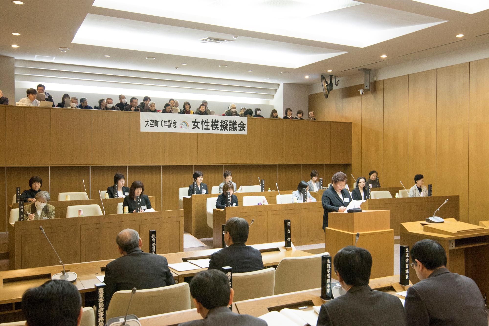 2階の席に座っているたくさんの傍聴者と発言台をはさむように座っているたくさんの模擬議員が見守る中、スーツを着た女性が会場の中央で発言をしている様子の写真