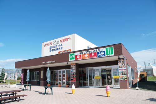表に施設案内が書かれ、屋外にソフトクリームの置物や飲食スペースが設けられた茶色い建物