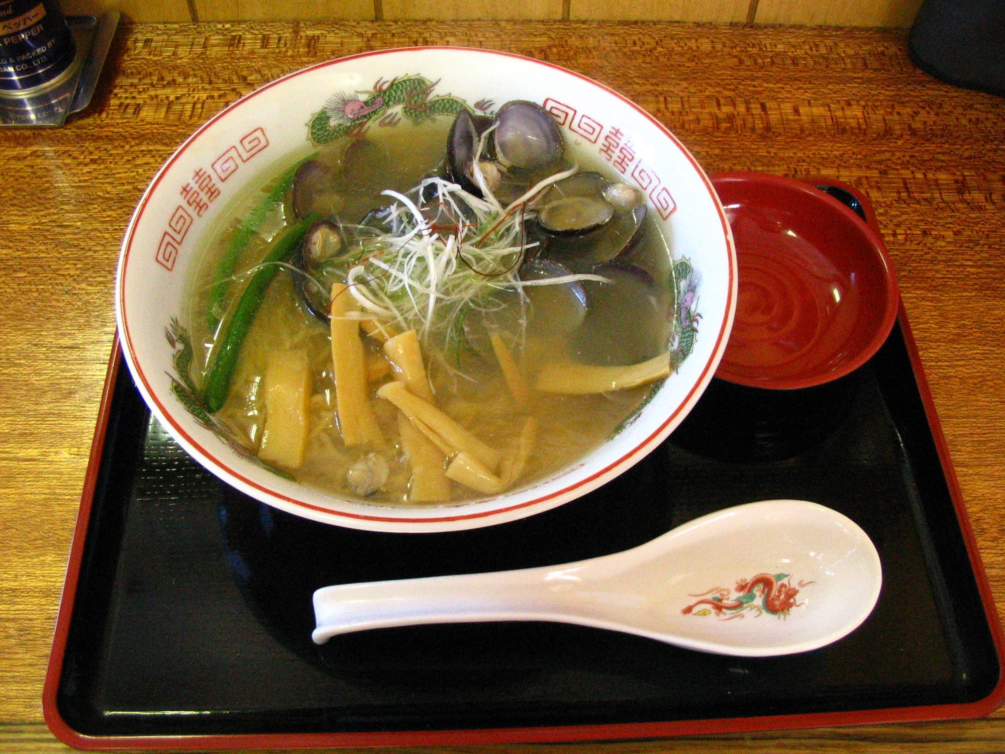 黒いトレーに沢山のシジミが入ったラーメンと赤い小皿とレンゲが置かれている写真