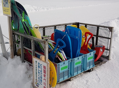 雪の上の網で囲われた場所に赤、青、黄色など様々な色や種類の貸し出し用ソリが置かれている写真