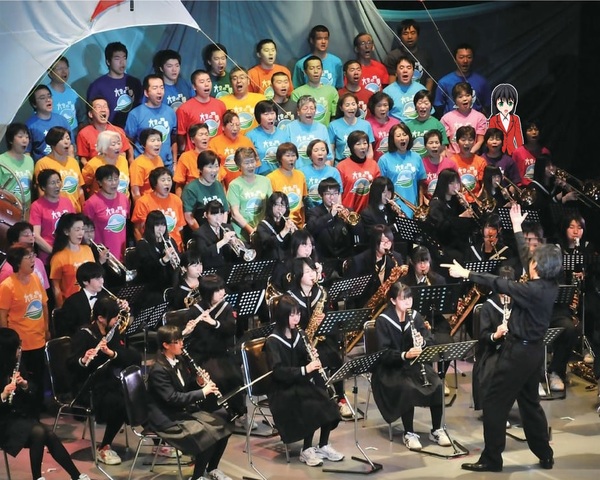制服姿のブラスバンドと色とりどりのTシャツ姿のコーラス隊が歌う合併5周年記念町民舞台の写真の中で一緒に歌う赤いジャケット姿の女の子のイラスト