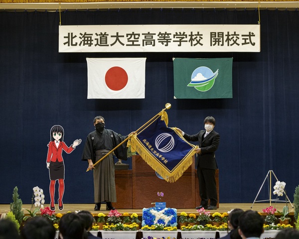 大空高等学校開校式ステージ上で校長が校旗を広げている写真の中に立ち左手で校旗を指し示す赤いジャケット姿の女の子のイラスト