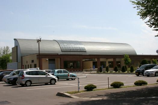 青空の下、大きな黒い扇形の屋根を持つ建物の手前の駐車場に数台の車が止まっている様子の写真