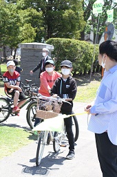 黄色い旗を上げている先生と自転車に乗って立ち止まっている小学生3人の様子の写真