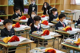 制服姿で椅子に座っている新一年生の机の上に黄色い通学帽や赤い体育帽などが置かれている写真