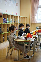 黄色い通学帽や赤い体育帽などが置かれている机の前で制服姿で椅子に座っている新一年生の3人の写真