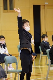 椅子から立ち上がって大きく右手を上げている新一年生の男の子の写真