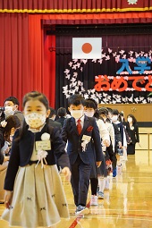 入学おめでとうと書かれた幕と日の丸を背に体育館を一列に並んで退場している新一年生たちの写真