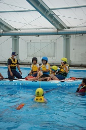 プールの救命ボートに乗っている救命胴衣をつけたスタッフの男性と4人の子どもたちのうちの1人がプールの中にいる子どもが持っている紐を引っ張っている様子の写真