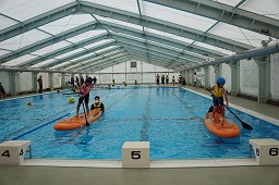 プールでそれぞれオレンジ色のカヌーの上に立ってパドルを動かしている男の子二人とカヌーを支えているスタッフの写真