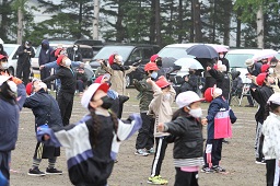 グラウンドで準備運動をしている紅白帽をかぶった小学生たちと傘をさしてそれを見ている父母の写真