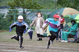 赤い帽子と白い帽子をかぶった男子小学生2人がグラウンドで走っている様子を見ている父母の写真