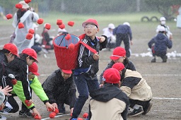 赤い帽子をかぶった小学生たちがひとりが背負っている赤いかごに赤い玉を入れている写真