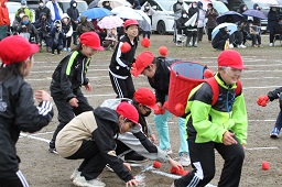 赤い帽子をかぶった小学生たちがひとりが背負っている赤いかごに赤い玉を入れている写真その2