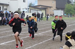 リレー競走で白いはちまきをして白いバトンを持った男子小学生を追いかけるそれぞれ黄色や赤のはちまきをしている小学生たちの写真