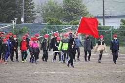 グラウンドで赤い旗を掲げている男子小学生と横二列になって更新している赤い帽子をかぶった小学生たちの写真