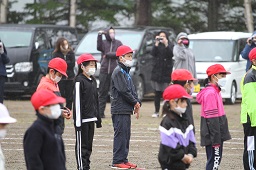 グラウンドで起立している赤い帽子の小学生たちとそれを撮影している父母たちの写真