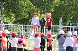 台に乗っている赤い帽子をかぶった3人の園児とそれぞれ赤い帽子や白い帽子をかぶっている園児たちの写真