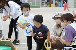 近距離にある輪投げの台に向かって青い輪っかを投げようとしている子どもとそれを見ている子どもと母親と先生の写真