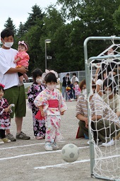 浴衣姿の女の子がサッカーボールを蹴ってゴールに入れようとしている様子を見守っている大人や子どもたちの写真