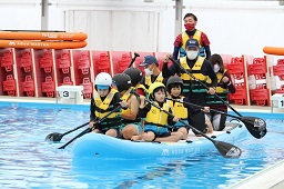 プールで子ども5人大人4人がひとつの青い救命ボートに乗ってパドルで漕いでいる様子の写真