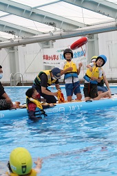 プールに浮いている男の子に向けてプール上の救命ボートから救命道具を投げている男の子とそれを見ているスタッフや子どもたちの写真