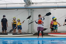 プールサイドで女性スタッフに合わせてパドルを漕ぐ練習をしている子どもたちの写真