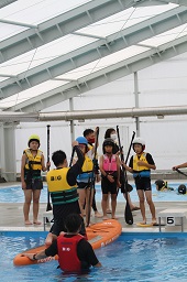 プール上でカヌーの漕ぎ方を教えているスタッフとその様子をプールサイドで見ている4人の子どもたちの写真