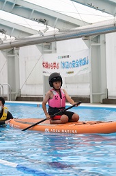 オレンジ色のカヌーの上に正座してパドルを漕いでいるピンクの救命胴衣を着た子どもの写真