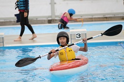 プールでカヌーに乗ってパドルを持っている黄色い救命胴衣と黒いヘルメットを付けた男の子の写真