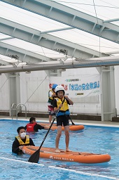 プール上でカヌーの上に立ってパドルを漕いでいる黄色い救命胴衣と白いヘルメットを付けた男の子とカヌーを支えているスタッフの写真